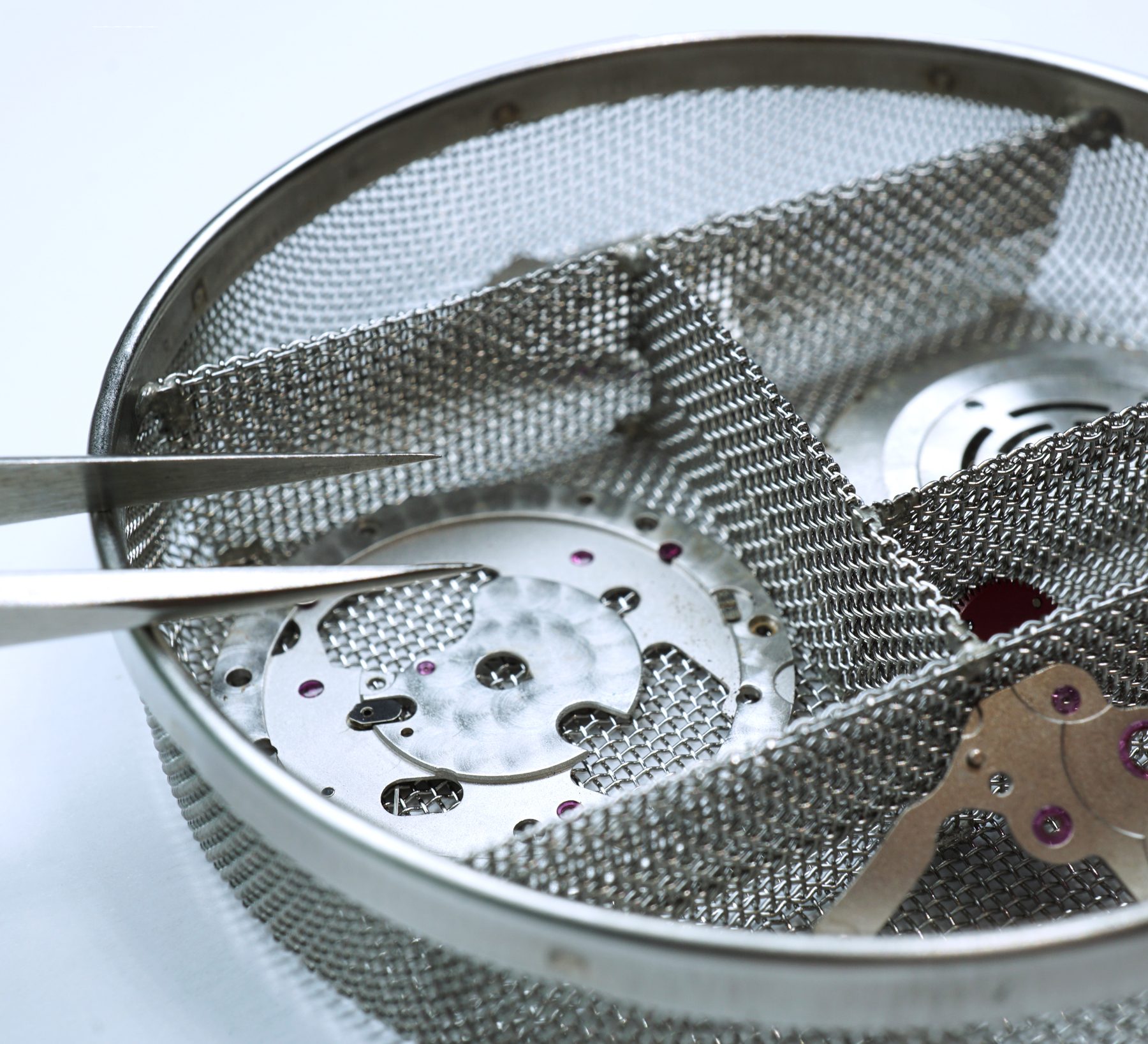 Watch movement components resting in a mesh cleaning basket, held with tweezers